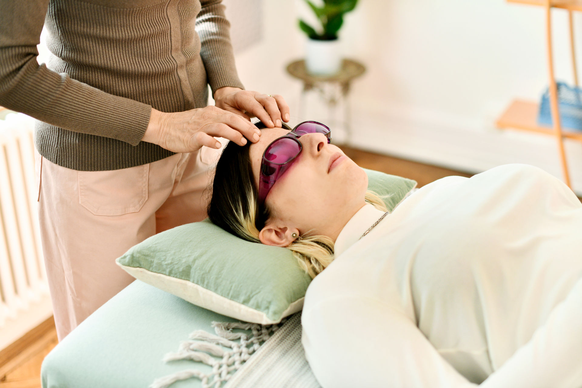 Behandlungssituation in der Kinesiologie-Praxis von Pascale Hauser. Eine Patientin liegt entspannt auf einer Liege, sie trägt eine lila Farbbrille. Pascale Hauser berührt die Stirn der Patientin mit den Fingern. Die Atmosphäre ist sehr entspannt.