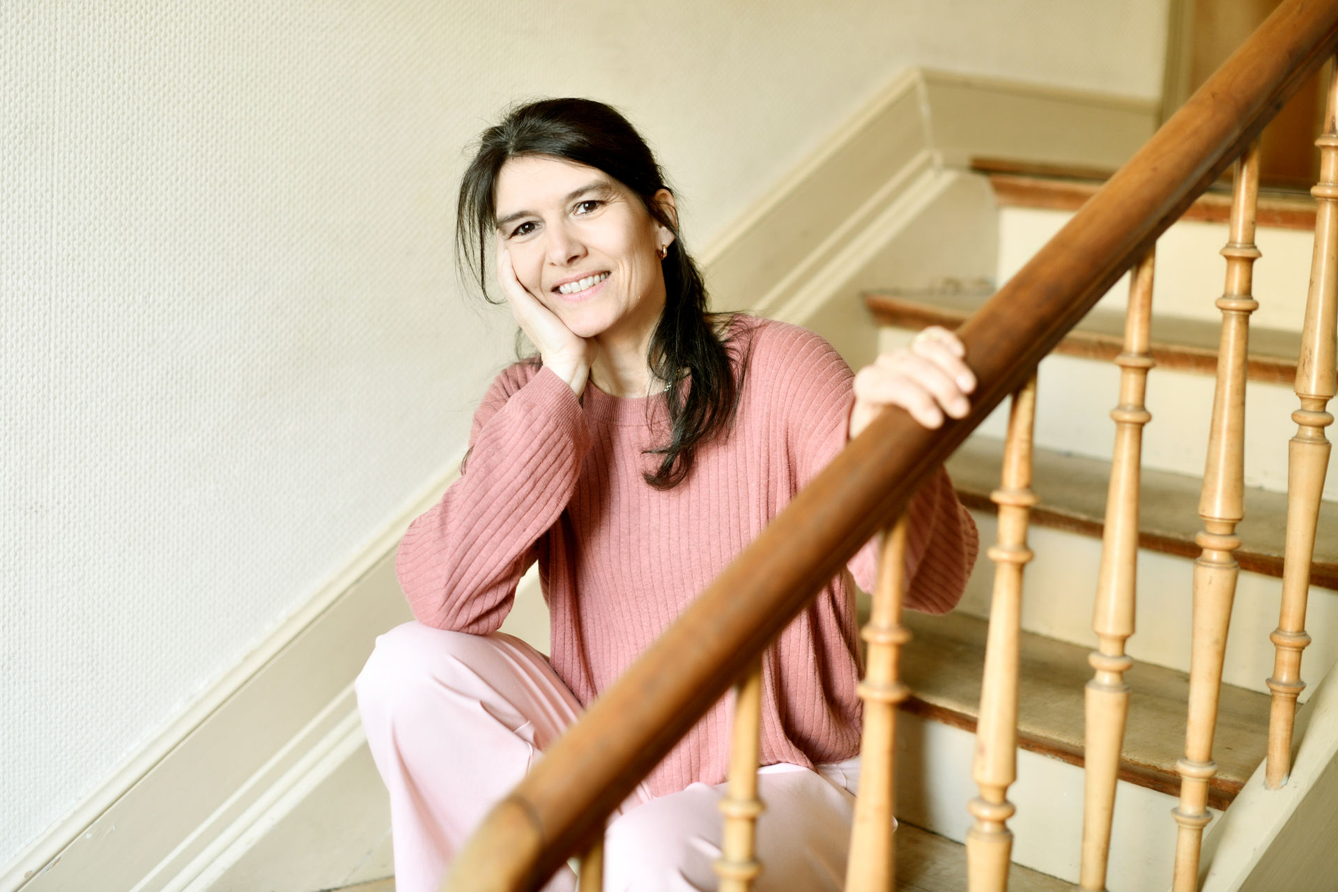 Portrait von der Kinesiologin Pascale Hauser. Sie sitzt auf der Treppe im Hausflur zur Praxis, der Kopf ist auf einer Hand abgestützt. Sie lächelt freundlich in die Kamera.