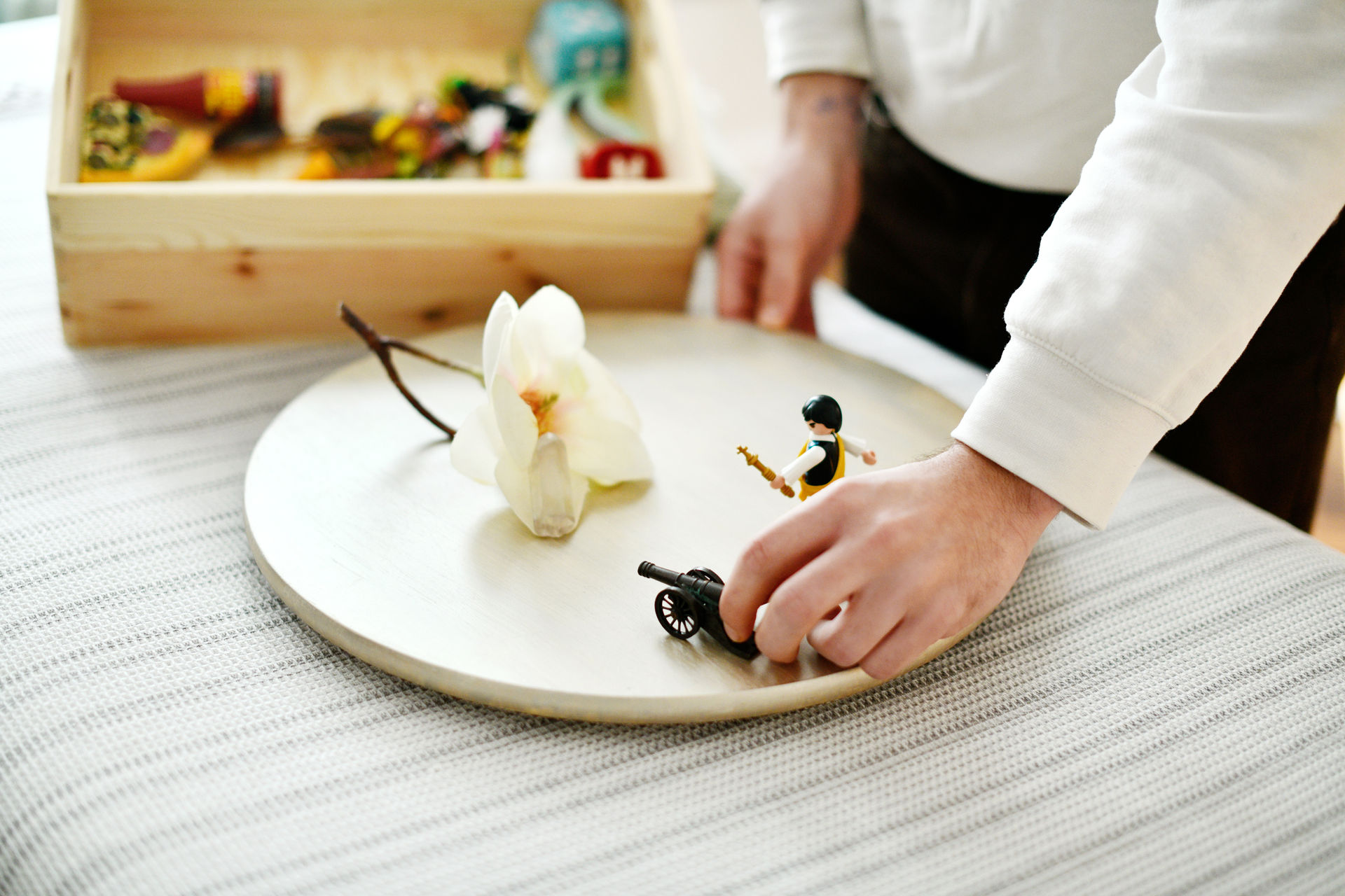 Systemische Aufstellung mit Figuren. Der Patient hat sich für eine Kunstblume, eine Playmobil-Kanone und ein Playmobilmännchen entschieden. Er stellt diese Dinge auf ein Holzbrett. Im Hintergrund ist unscharf eine Kiste mit weiteren Gegenständen zu erkennen.